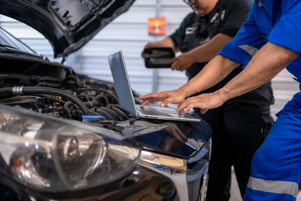Photographie d'un atelier automobile professionnel montrant une équipe de 3 à 4 techniciens en tenue de travail (bleu de travail, gilets) travaillant à différentes stations : un mécanicien sous un véhicule surélevé, un carrossier en atelier de peinture en arrière-plan, un électricien inspectant un système hybride, tous concentrés sur leurs tâches respectivas. L'atelier doit être moderne, bien équipé avec pont élévateur, équipements de diagnostic, outils professionnels visibles, logo ou enseigne du garage visible mais discrète. Atmosphère professionnelle, collaborateurs engagés dans leur spécialité respective, démontrant la pluridisciplinarité et l'expertise de l'équipe.